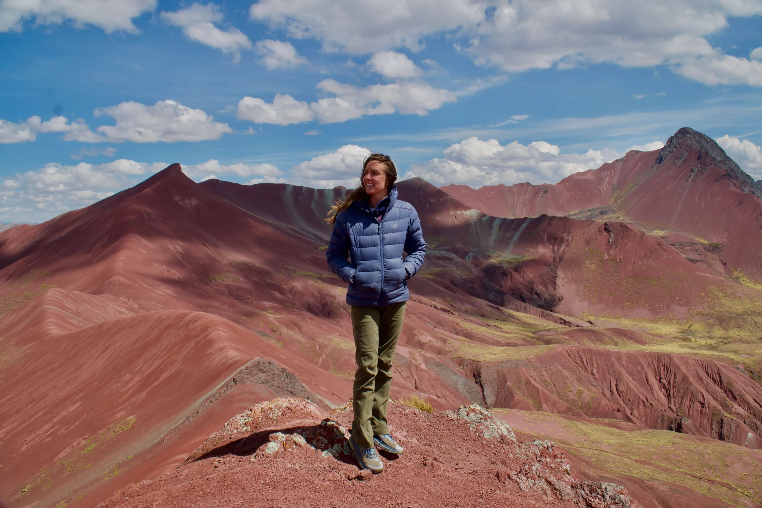 solo female traveler standing on red rock overlooking red uneven landscape rainbow mountain peru